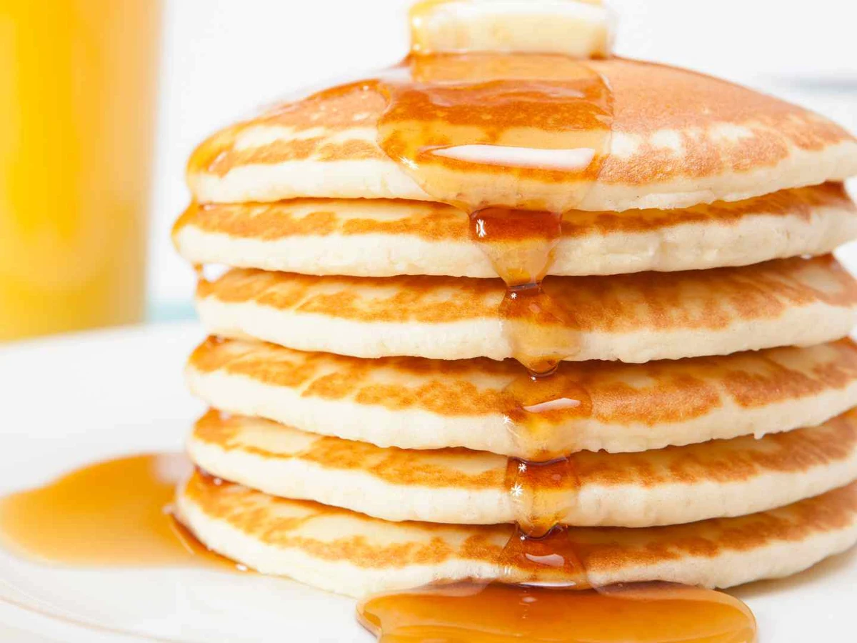 A tall stack of pancakes with fruit, syrup, and a dish of cream.