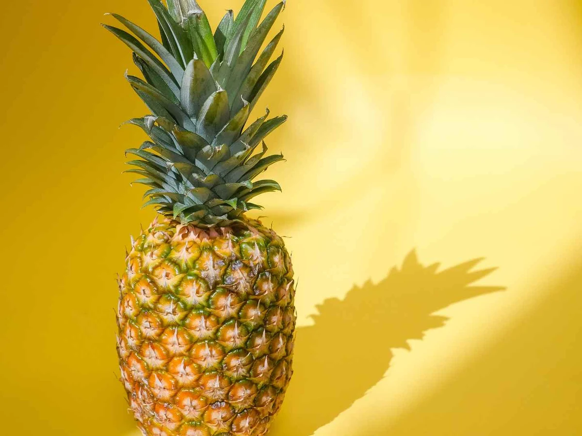 A whole pineapple standing against a bright yellow wall and casting a shadow.