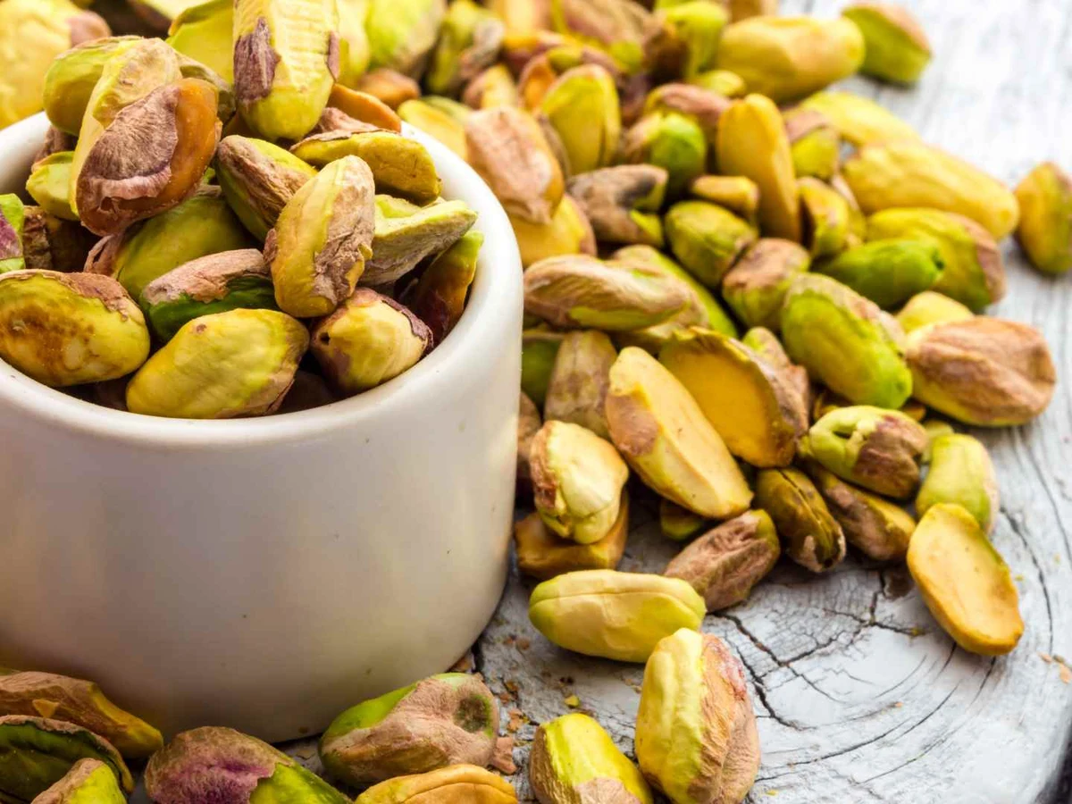 Shelled pistachio kernels fill a white cup and cover the surrounding surface.