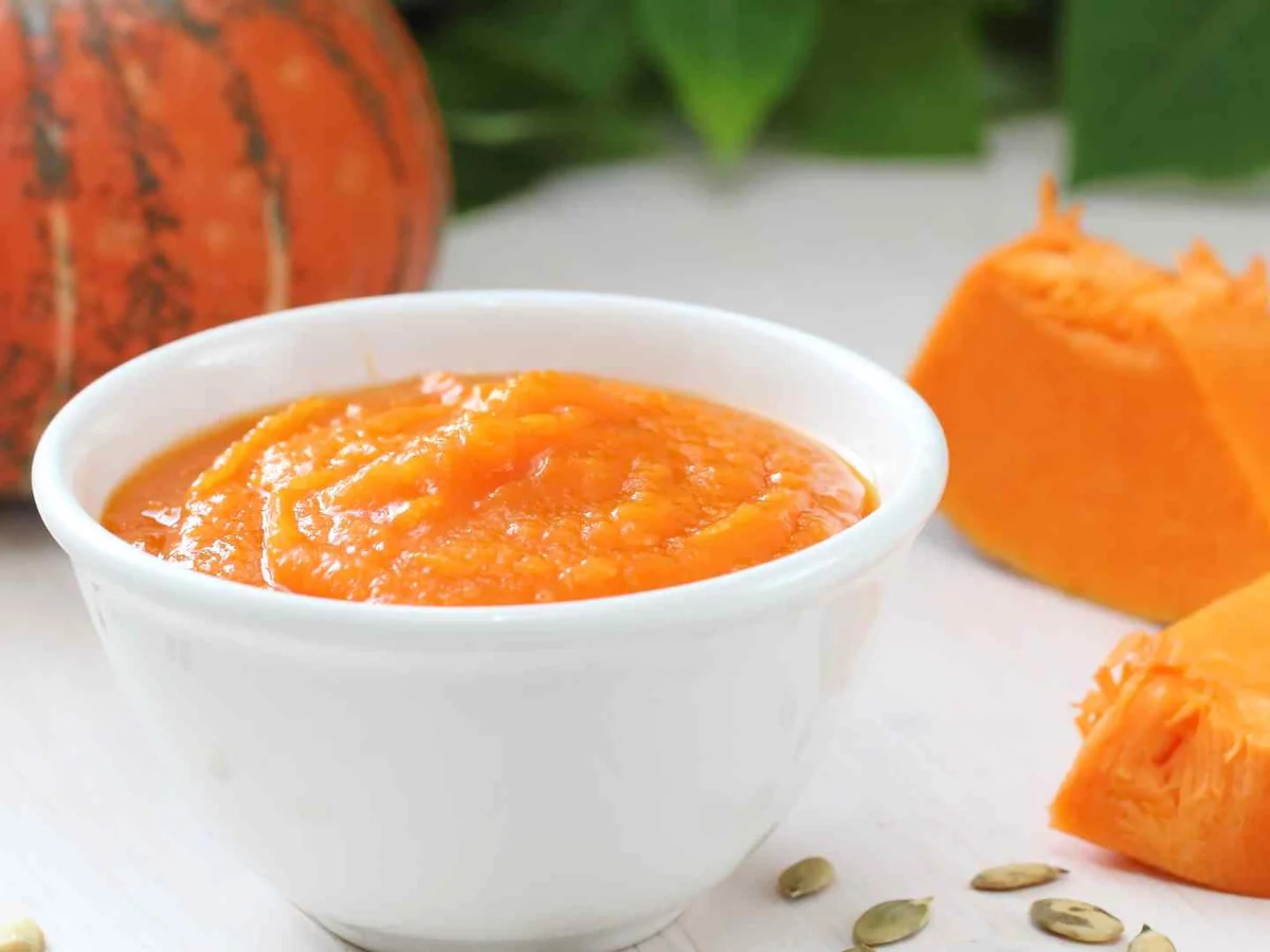 A white bowl of pumpkin puree with pumpkin and seeds behind it