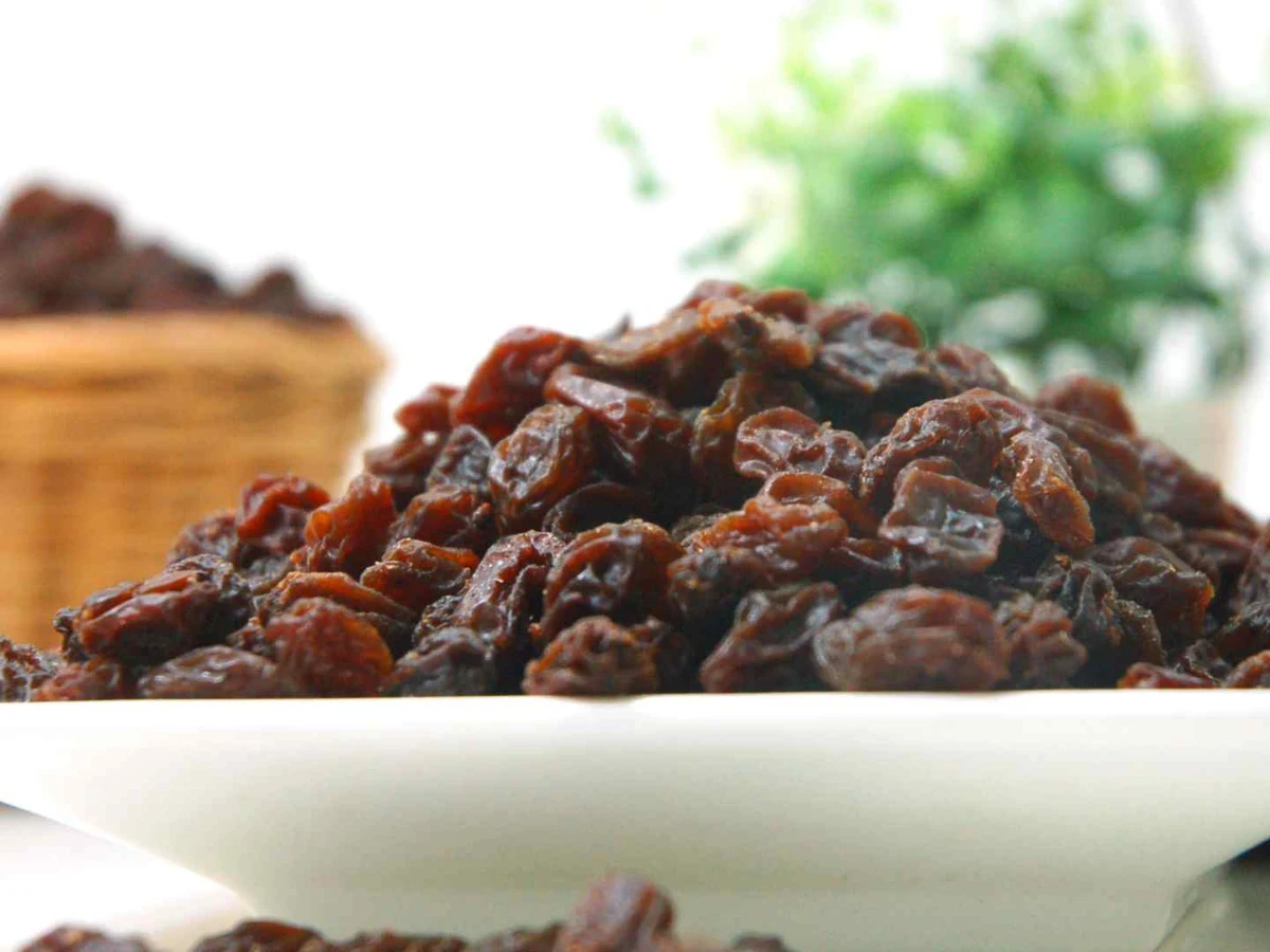 Dark brown raisins heaped on a white plate with a wicker basket behind it.