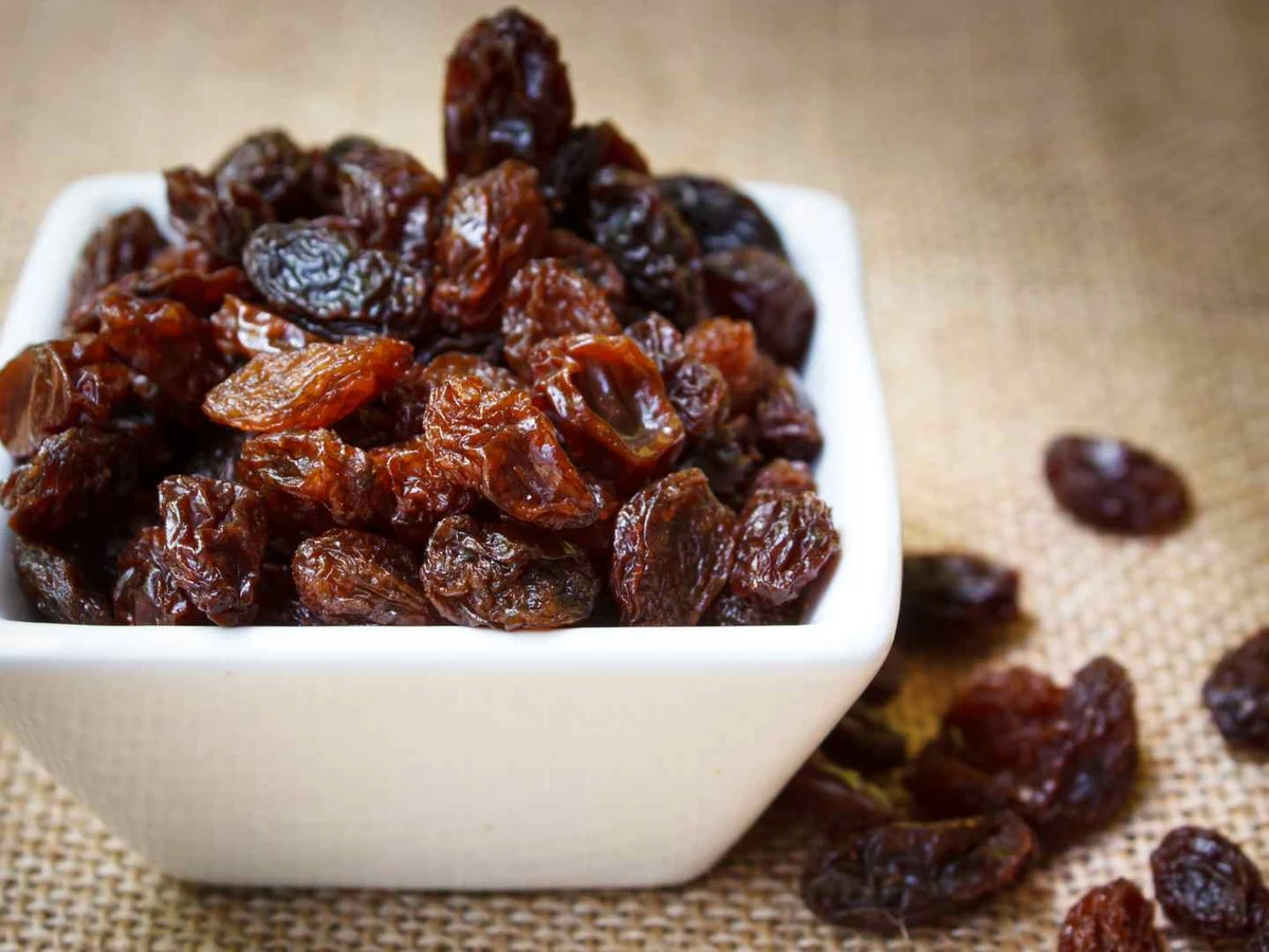 Dark brown raisins in a square white bowl with loose raisins on burlap.