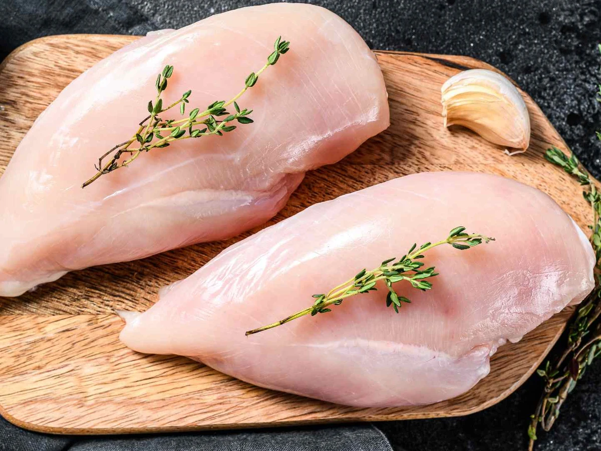 Two raw boneless chicken breasts on a wooden board with garlic and herbs.