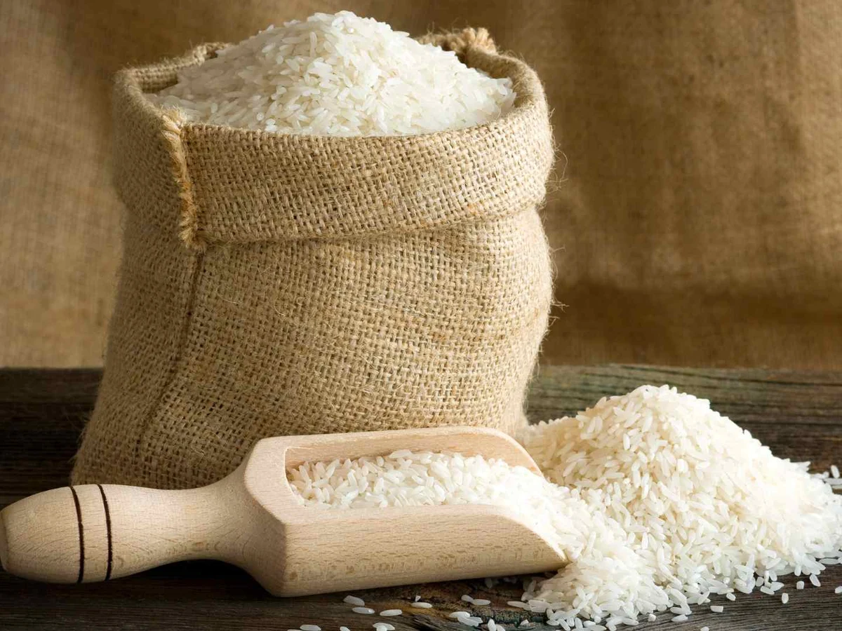 Uncooked white rice in a burlap sack beside a wooden scoop and small pile.
