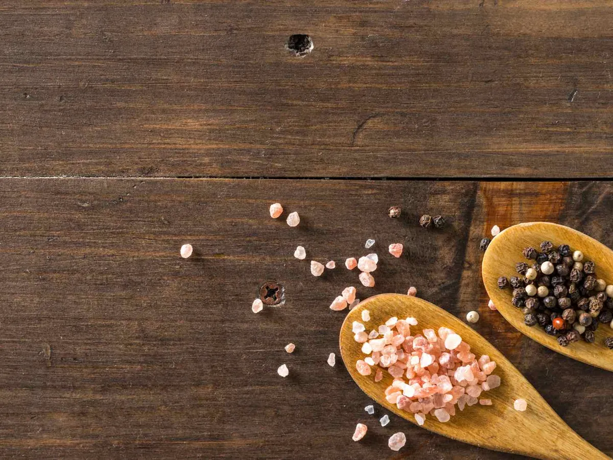 Two wooden spoons holding pink salt and black pepper on a wood surface