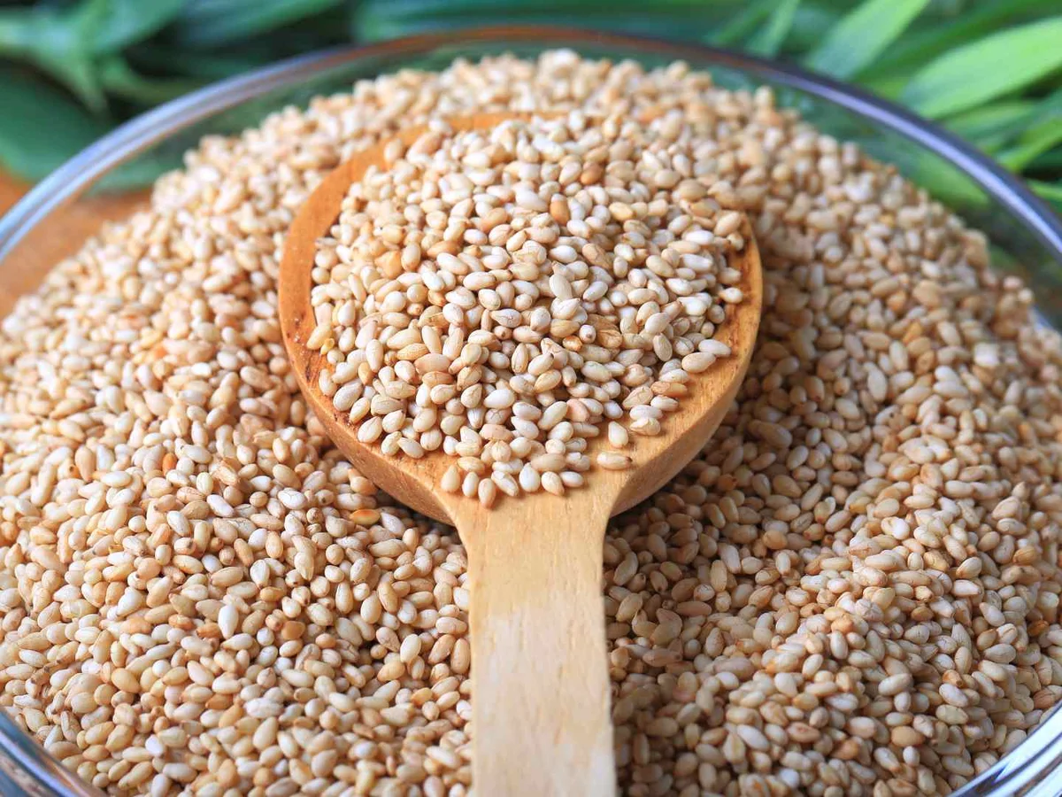 White sesame seeds piled in a glass bowl with a wooden spoon.