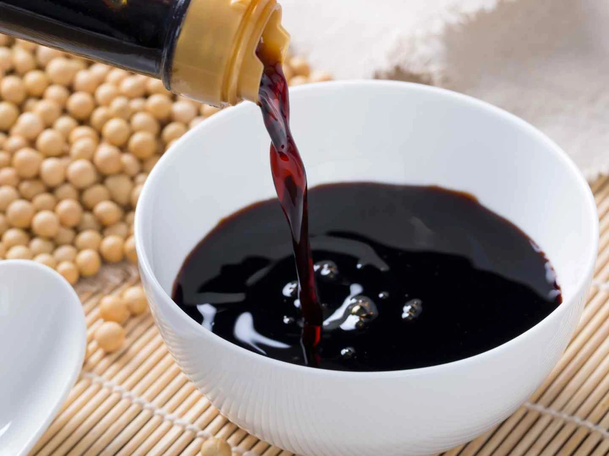 Dark soy sauce being poured into a white bowl beside soybeans.