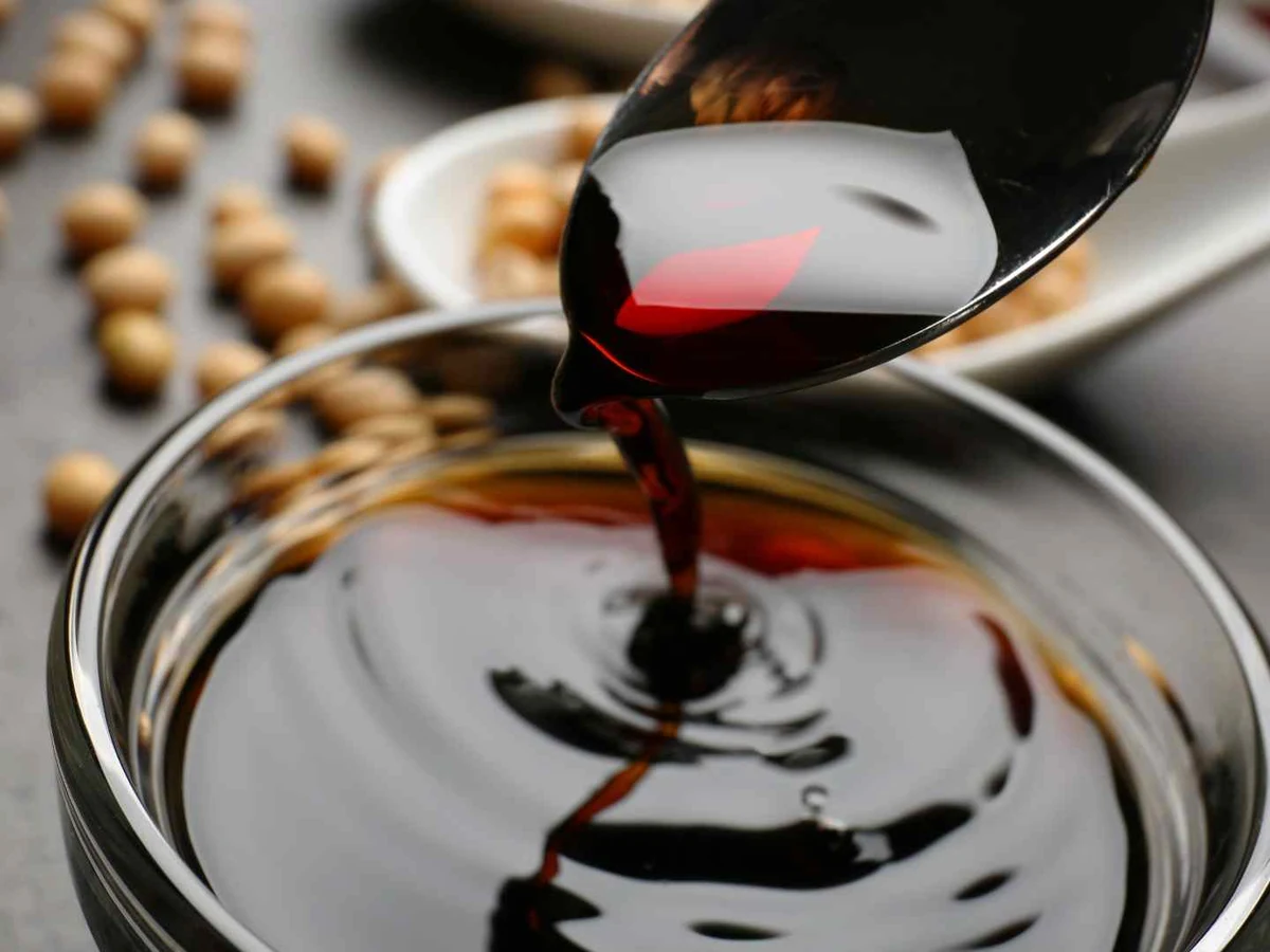 Dark soy sauce pouring from a spoon into a clear bowl.