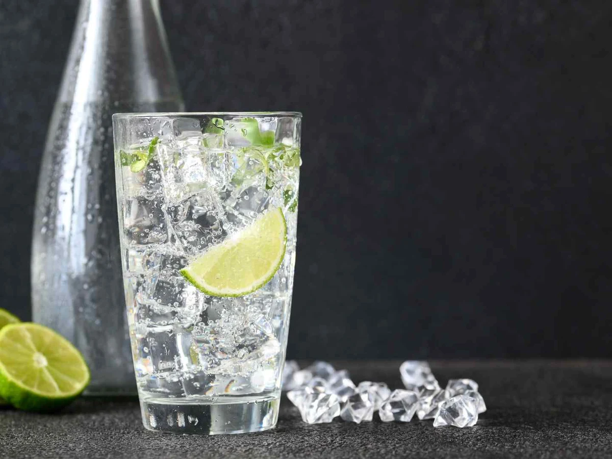 Sparkling water with lime in a glass beside a clear bottle and ice.