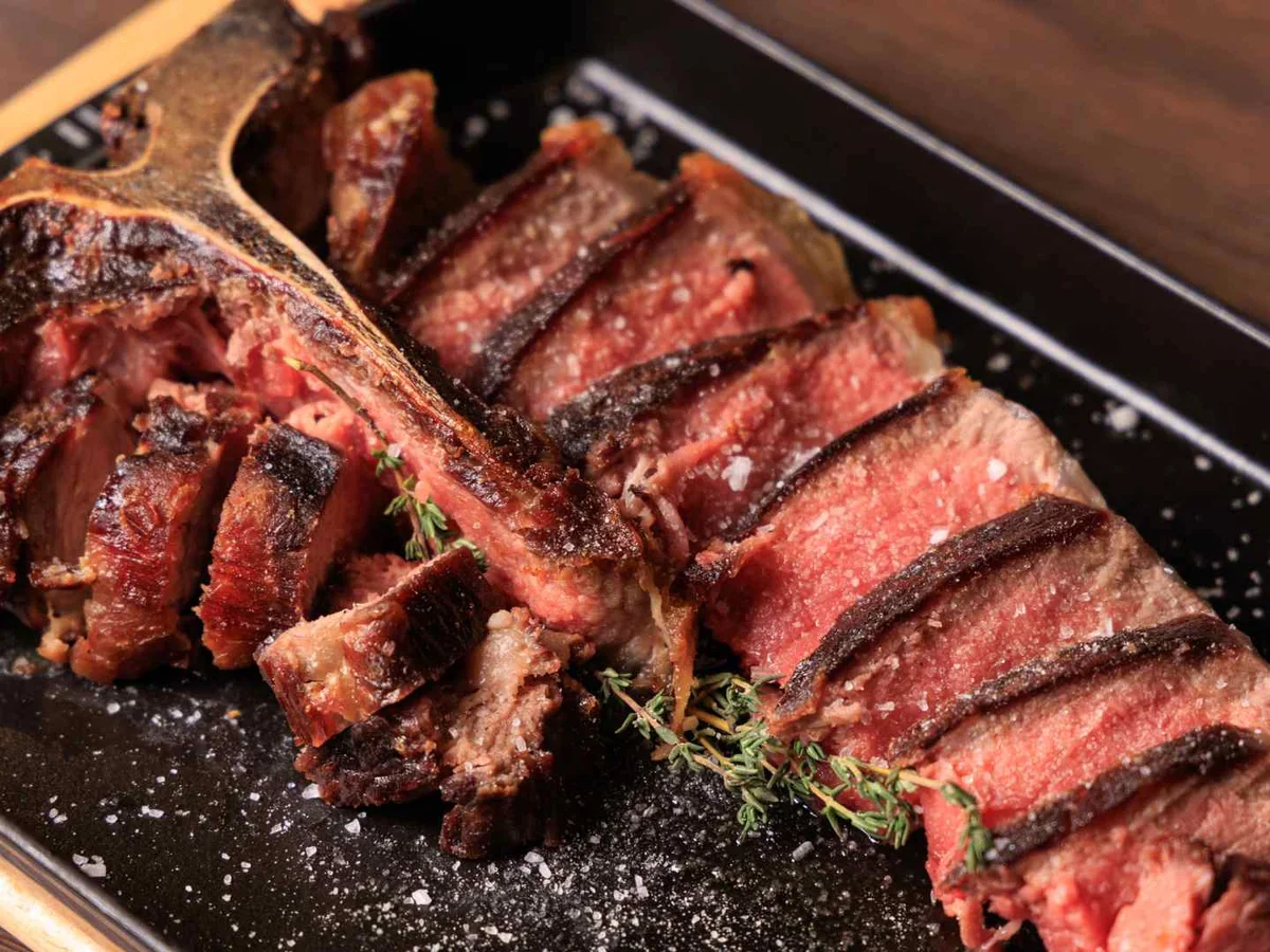 A sliced grilled bone-in steak on a dark metal tray.