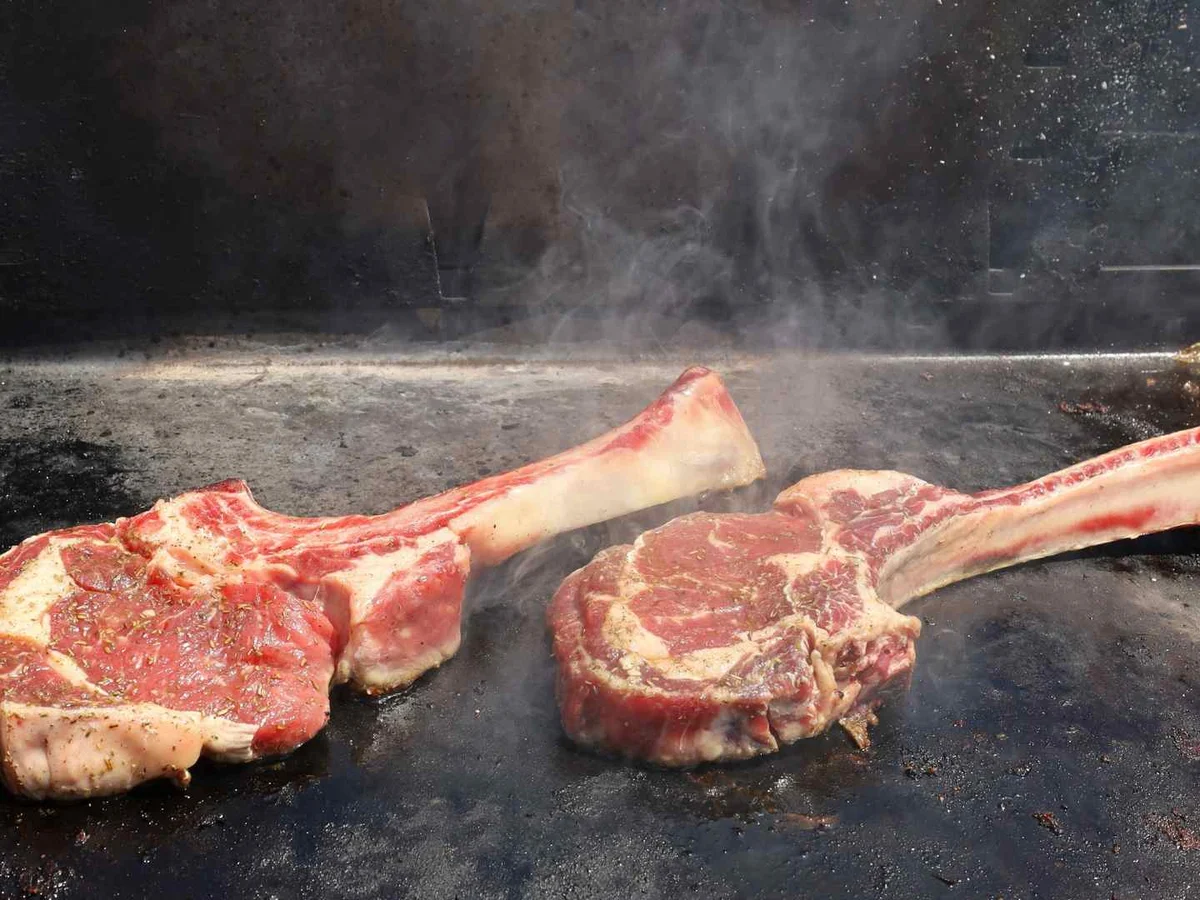 Two raw bone-in steaks searing in a black pan with visible steam.