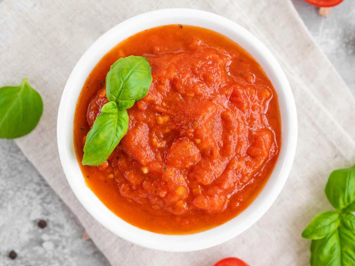 Tomato sauce in a white bowl garnished with basil on a pale table.