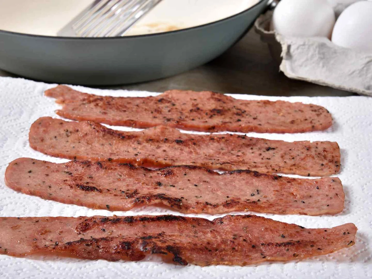 Cooked turkey bacon strips draining beside eggs and a pan