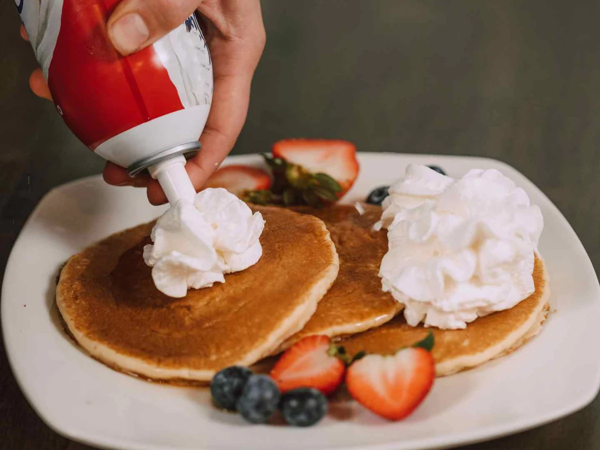 A hand dispensing whipped cream onto pancakes topped with fruit