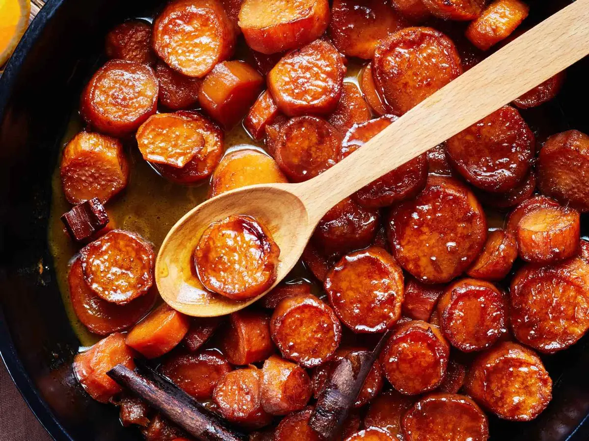 Glazed orange yam rounds in a dark pan with a wooden spoon.