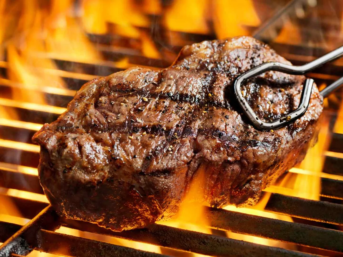 A thick steak held over orange flames on a grill