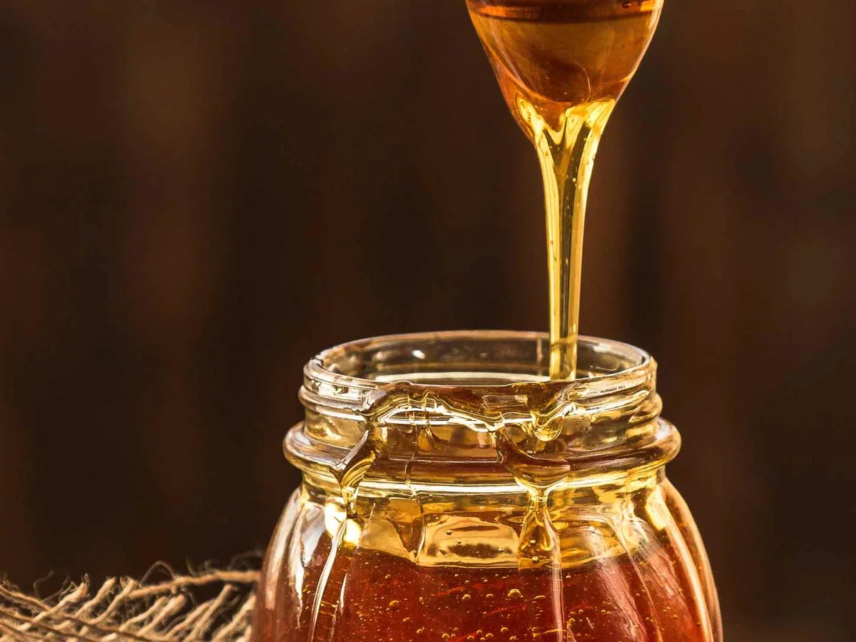 Golden honey pouring from a dipper into a glass jar