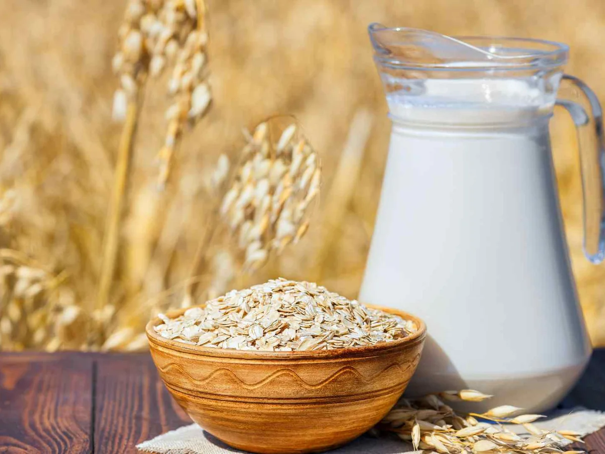 A glass pitcher of oat milk beside oats and wheat stalks