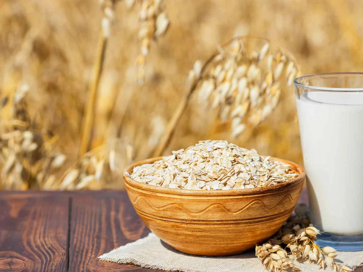 A glass of oat milk beside a bowl of oats and wheat stalks