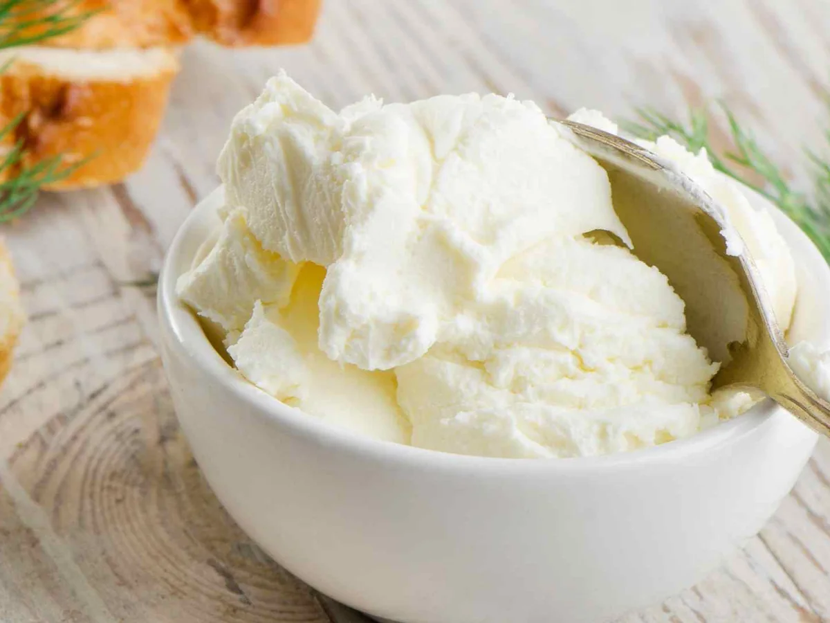 Cream cheese in a white bowl beside bread and herbs