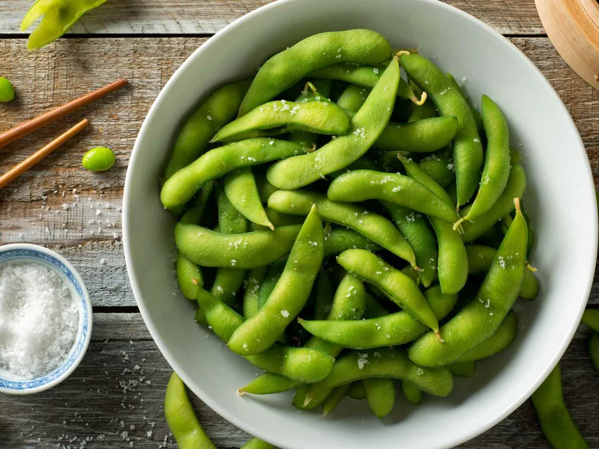 Edamame pods in a white bowl beside chopsticks and salt