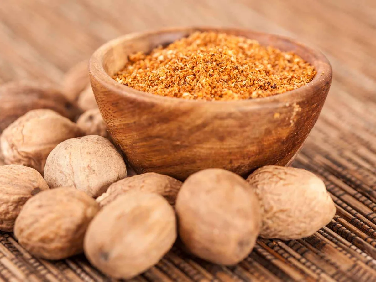 Ground nutmeg in a wooden bowl beside whole nutmegs