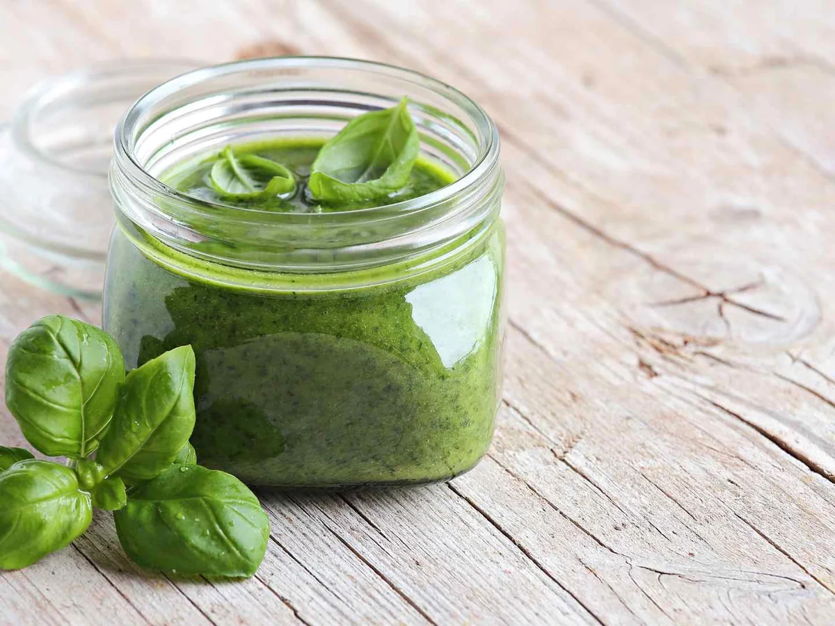 A glass jar of green pesto with fresh basil leaves