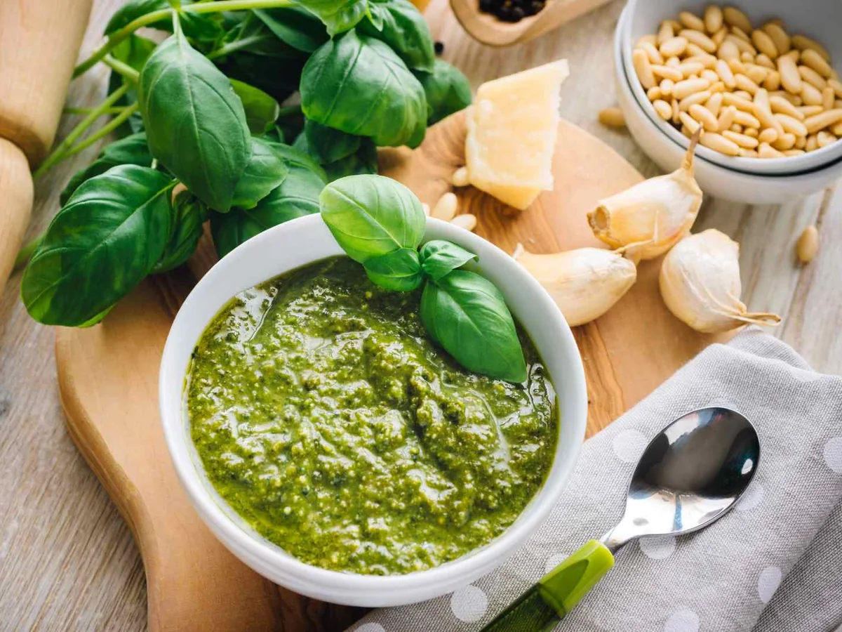 A bowl of pesto surrounded by basil, pine nuts, and cheese
